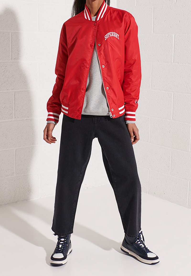 Personne debout contre un mur de briques blanches portant une veste rouge Superdry, un t-shirt gris, un pantalon noir et des baskets noires et blanches.