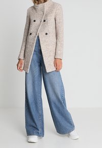 Cappotto doppiopetto in misto lana beige con bavero alto e bottoni neri, abbinato a jeans in denim blu a gamba larga e sneakers bianche.