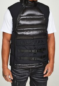 Mann, der eine schwarze gepolsterte Weste mit dem Schriftzug "GLORIOUS" und Schnallenriemen über einem weißen T-Shirt mit kurzen Ärmeln und dunklen Jeans trägt, mit tätowierter Hand.