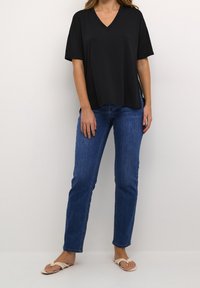 T-shirt nera a V con maniche corte, vestibilità rilassata. Indossata con jeans blu e sandali beige, presenta un design minimalista e un look informale.