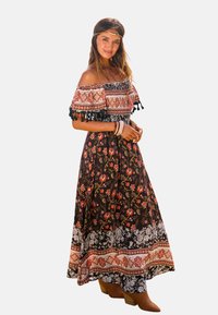 Abito maxi off-the-shoulder in nero con motivi floreali, accentuato da un corpetto patterned e maniche frangiate, abbinato a stivali color sabbia.