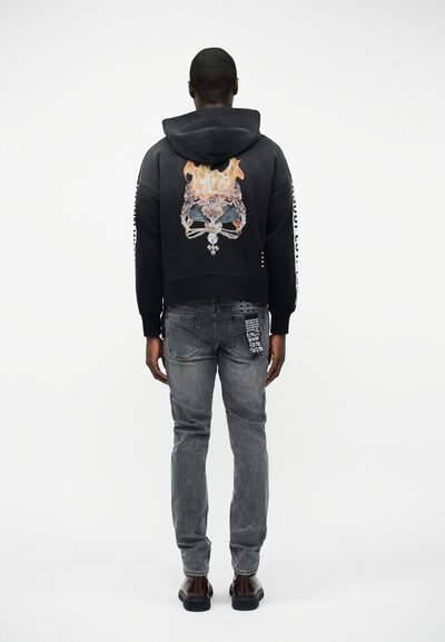 Άνδρας που φοράει μαύρο φούτερ με κουκούλα με γραφικό φλεγόμενης κορώνας στην πλάτη, γκρι τζιν με μπαλώματα στην πίσω τσέπη και σκούρα παπούτσια, στέκεται με την πλάτη στραμμένη.