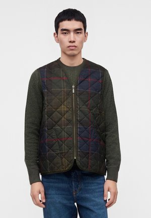 Jeune homme portant une veste matelassée à carreaux vert foncé sur un pull vert et un jean bleu, debout devant un fond clair uni.