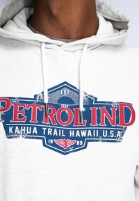 Sudadera blanca con un diseño gráfico desgastado en azul y rojo que incluye el texto "PETROL IND" y "KAHUA TRAIL HAWAII U.S.A." Capucha ajustable con cordón.