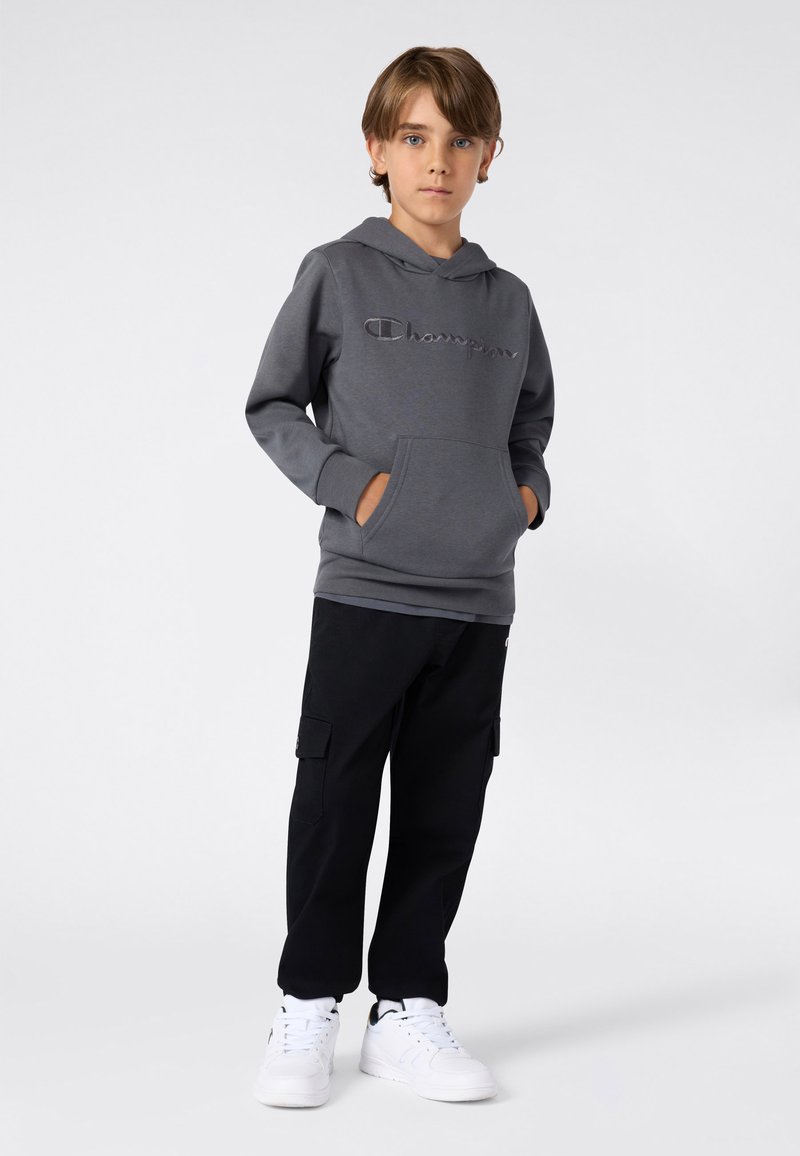 Ragazzo in piedi con le mani in tasca, indossa una felpa grigia Champion, pantaloni cargo neri e sneakers bianche, su uno sfondo chiaro e semplice.