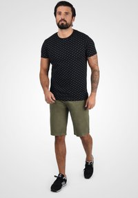 Sort t-shirt med hvide prikker, parret med olivengrønne shorts og sorte træningssko. T-shirten har rund halsudskæring og korte ærmer.