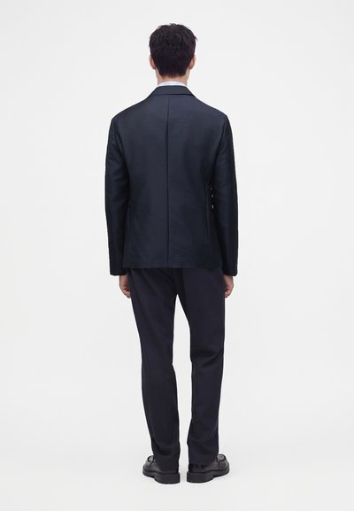 Homme portant une veste de costume noire, un pantalon foncé et des chaussures noires, debout de dos devant un fond blanc uni.