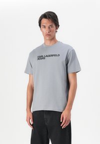Pelēks kokvilnas T-krekls ar īsām piedurknēm un apaļu kakla izgriezumu. Priekšpusē redzams teksts "KARL LAGERFELD JEANS" treknrakstā, melnā krāsā.