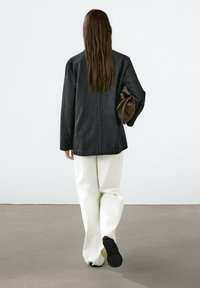 Veste en denim foncé à la coupe décontractée, associée à un pantalon blanc à jambes larges. Pochette en cuir portée sous un bras, chaussures noires discrètes.