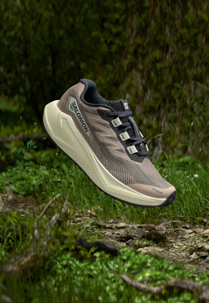 Beige en zwarte Salomon-hardloopschoen zwevend boven een met gras en stenen bedekte bosbodem in de buitenlucht.