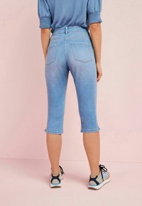 Pantalons capri en denim bleu clair avec taille haute, ourlets retroussés et deux poches au dos, associés à des baskets légères avec des accents noirs, bleus et beiges.