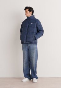 Chaqueta de plumas azul marino con diseño acolchado y cuello alto, combinada con jeans de pierna ancha en azul claro y zapatillas blancas. Marca mínima visible.
