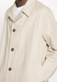 Beiger Mantel mit Knöpfen und weitem Kragen, getragen über einem weißen Hemd, mit detaillierter Nahtführung und seitlichen Taschen, wobei eine Hand in der Tasche steckt.