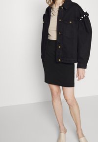 Chaqueta de mezclilla negra con botones dorados, dos bolsillos frontales y mangas abullonadas adornadas con perlas blancas, combinada con una falda negra y zapatos beige.