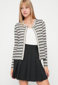 Stripete cardigan i krem og svart, ribbestrikket tekstur, rund halslinning, knappestengning, parret med en mørk plisserte skjørt.