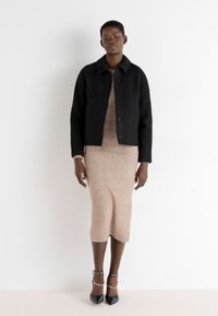 Black wool blend jacket with a collar, paired with a fitted beige ribbed dress. Model wears black, pointed-toe heels with ankle straps.