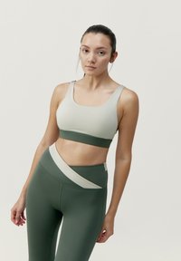 Reggiseno sportivo in verde chiaro e verde scuro, realizzato in tessuto elastico e liscio, presenta una fascia ampia sotto il busto e spalline spesse. I leggings hanno un design angolato con pannelli di colore contrastante.