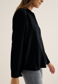 Chemise noire à manches longues avec un col, texture lisse, et coupe décontractée, portée sur un jean gris clair.