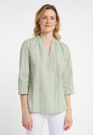 Femme portant un chemisier à rayures verticales vertes et blanches avec des manches trois-quarts et un pantalon blanc sur un fond blanc uni.