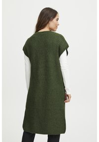 Olíva zöld kötött ujjatlan mellény texturált mintázattal, lekerekített nyakkivágással és oldalsó részekkel a szabad mozgásért. Fehér hosszú ujjú felsőre van felvéve.