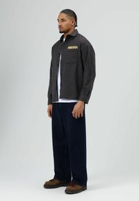 Zwart geruit overhemd met de tekst "FREE BOOM", gedragen over een wit T-shirt. Blauwe broek en bruine laarzen maken de outfit compleet.