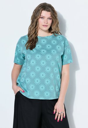 Femme aux cheveux ondulés portant un t-shirt manches courtes couleur sarcelle avec des motifs de soleil et un pantalon noir, debout contre un mur blanc.