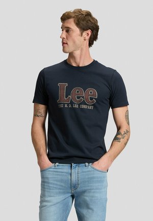 Uomo con capelli castani corti, maglietta blu navy "Lee" e jeans azzurri, sta in piedi con le mani in tasca e guarda alla sua sinistra. Tatuaggi sulle braccia.