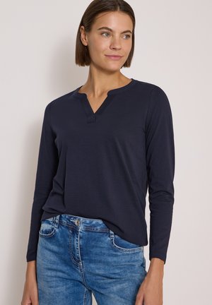Jeune femme aux cheveux bruns et raides, portant un haut bleu marine à manches longues et un jean bleu, regardant légèrement sur le côté.