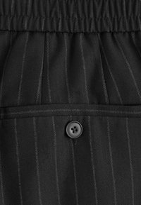 Tessuto nero con un motivo a spina di pesce e sottili righe sottili. Include una cintura elastica e un dettaglio con bottone nero.