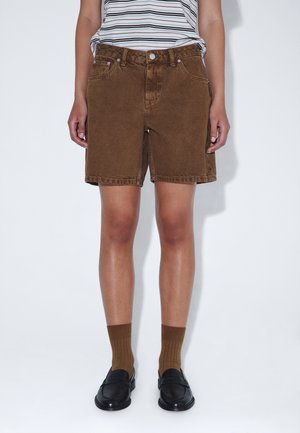 Persoon die bruine denim shorts, bruine sokken, zwarte loafers en een wit-zwart gestreept shirt draagt, staand tegen een witte achtergrond.