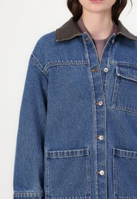 Denim jakna v modri barvi s rjavim ovratnikom, z gumbom na sprednji strani, dvema sprednjima žepoma in teksturirano tkanino.