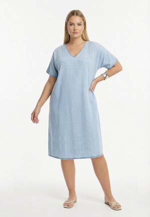 Kvinde iført en lyseblå knælængde denimkjole med korte ærmer og udsmykket halsudskæring, kombineret med beige sandaler og sølvsmykker.