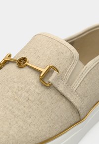 Chaussure à enfiler en tissu beige avec semelle blanche et détail décoratif en métal doré sur le dessus.