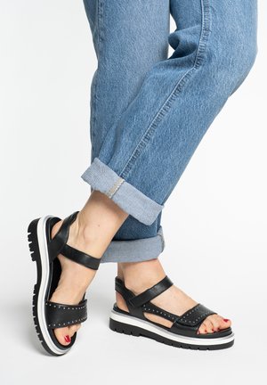 Feet in black studded sandals with thick white-soled platform, paired with cuffed blue jeans against a plain white background.