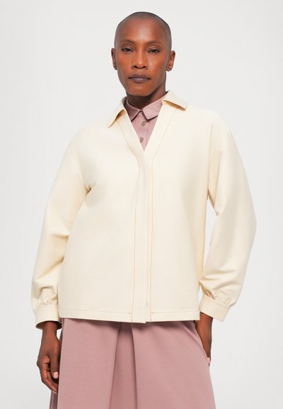 Κρέμα μακρυμάνικο μπλουζάκι με κολάρο και V-λαιμόκοψη, με αντιθετικά ραφάκια και χαλαρή γραμμή, συνδυασμένο με ροζ πουκάμισο από κάτω.