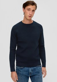 Granatowy sweter z okrągłym dekoltem i prążkowanym wzorem, o teksturowanej tkaninie, z rękawami raglanowymi i dopasowanymi mankietami, w zestawie z jasnoniebieskimi dżinsami.