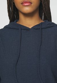 Primo piano di una persona con capelli intrecciati che indossa orecchini a cerchio in argento e una felpa blu navy a coste con cordini.