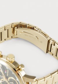 Montre-bracelet en acier inoxydable doré avec un bracelet en maillons métalliques, montrant le mécanisme du fermoir et une partie du cadran avec des boutons.