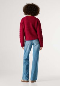 Raudonas megztinis su tekstūruotais, pintiniais rankovėmis, derinamas su šviesiai mėlynais, platėjančiais džinsais. Apranga pasižymi laisvu siluetu ir ribiniu apačiu.