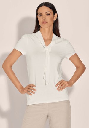 Vrouw met lang donker haar, die een wit topje met korte mouwen en een strikdetail aan de voorkant draagt en beige broek, staand met handen in de zij.