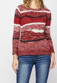Rot und weiß gestreifter Strickpullover mit wellenförmigen Mustern, langen Ärmeln, geripptem Saum und kontrastierenden schwarzen Akzenten. Getragen zu blauen Jeans.
