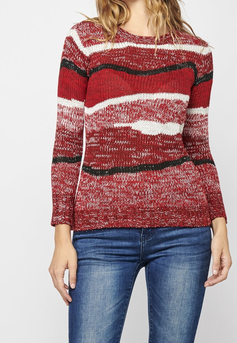 Rot und weiß gestreifter Strickpullover mit wellenförmigen Mustern, langen Ärmeln, geripptem Saum und kontrastierenden schwarzen Akzenten. Getragen zu blauen Jeans.
