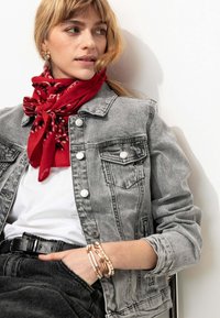 Veste en denim grise avec des boutons argentés, chemise blanche en dessous, écharpe à motifs rouges, ceinture noire et bracelets superposés au poignet.
