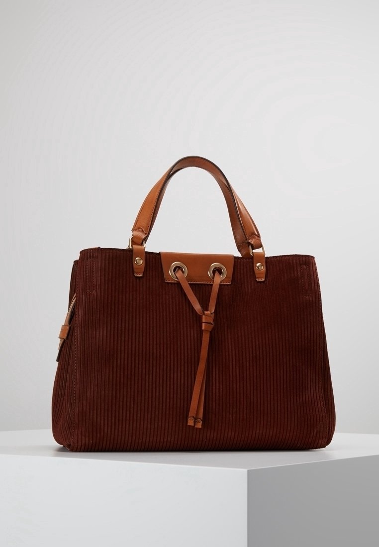 Sac à main en velours côtelé marron avec anses en cuir camel et détail à cordon, placé sur un piédestal blanc sur un fond clair uni.