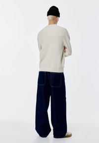 Beiger gerippter Pullover, lockere Passform, kombiniert mit weit geschnittenen dunkelblauen Jeans. Eine schwarze Strickmütze und helle Schuhe vollenden den Look.