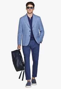 Mann trägt blau kariertes Sakko, marineblaues Poloshirt, marineblaue Hose, blaue Loafers, Sonnenbrille, trägt einen dunkelblauen Rucksack in der rechten Hand und geht vorwärts.