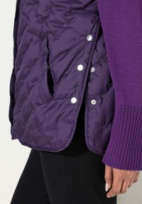 Lila Puffer-Weste mit gesteppter Textur, Druckknöpfen und seitlichen Taschen, kombiniert mit einem lila gerippten Pullover und schwarzen Hosen.