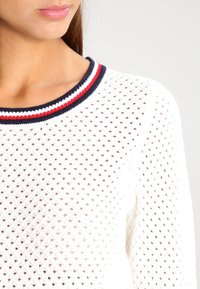 Maglione bianco lavorato a maglia con un tessuto strutturato e traspirante. Presenta un collo a coste con strisce blu navy, rosse e bianche. Primo piano dell'area del collo.