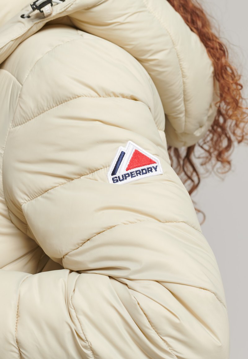 Giacca imbottita color crema con un design trapuntato e una finitura testurizzata. Include un patch con il logo rosso, bianco e blu.