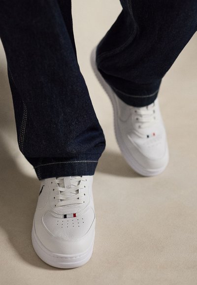 Λευκά δερμάτινα αθλητικά παπούτσια με κόκκινες, μαύρες και λευκές λεπτομέρειες, φορεμένα με σκούρο μπλε τζιν σε ανοιχτό γκρι πάτωμα.